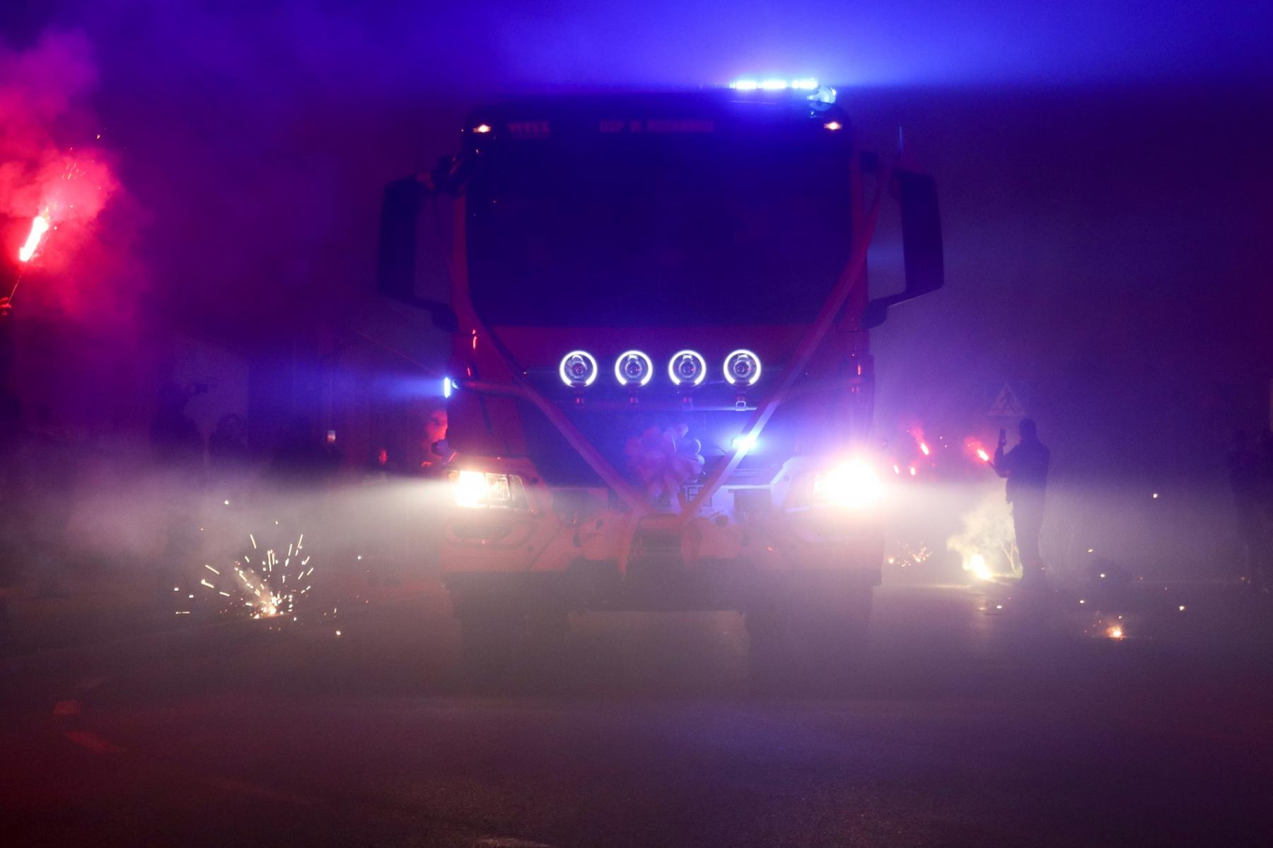 wóz strażacki w zmroku oświetlony, po bokach  palą się race,  