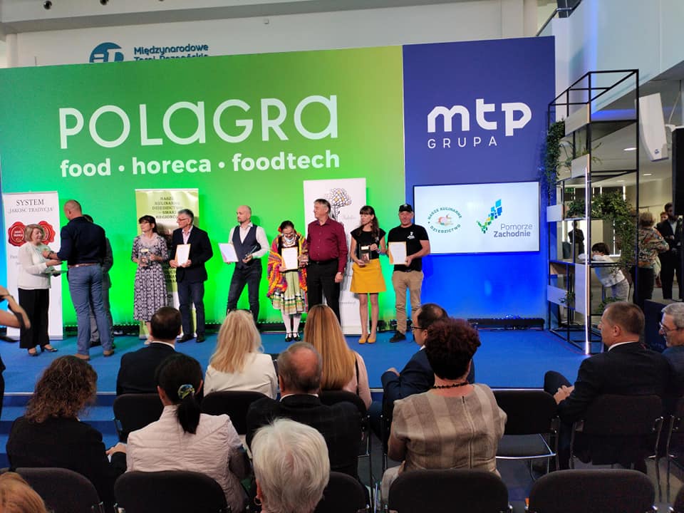 Zdjęcie przedstawia osoby w oddali na scenie. Pierwsi od prawej stoją państwo Dobropolscy.Z tyłu plansze zielone i niebieskie. Na zielonej duży napis POLAGRA